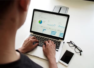 Person sitzt vor einem Laptop auf dem Statistiken zu sehen. Nebendran ein Handy und eine Brille. 